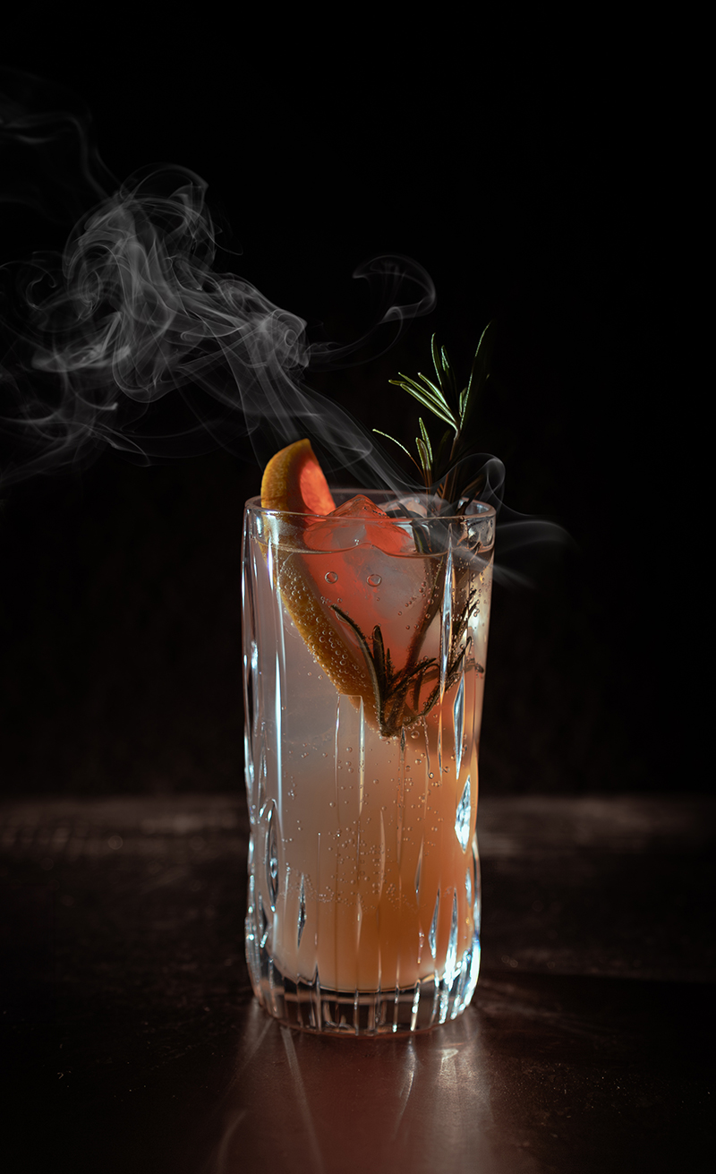 Afbeelding van een Paloma cocktail op mysterieus donkere achtergrond. Er komt rook uit het cocktailglas met garnituren als rozemarijn en een stukje pompelmoes.
