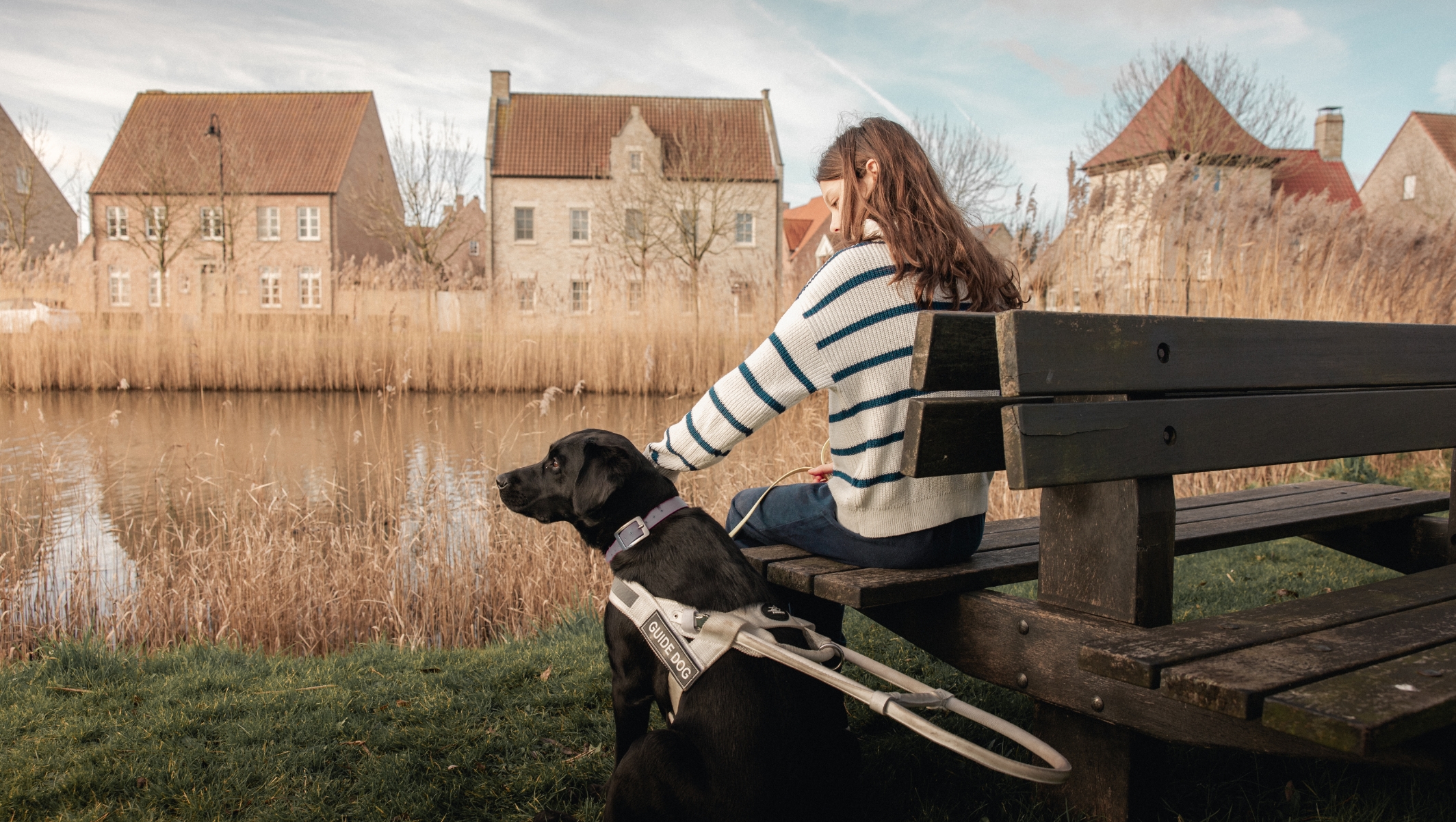 Afbeelding van een jong meisje dat op een houten bank zit naast haar blindengeleidehond. Ze bevinden zich voor een meer met hoge grassen en huizen op de achtergrond. Het meisje aait haar hond die een harnas draagt. We zien de rug van het meisje en de hond die genieten van het moment.