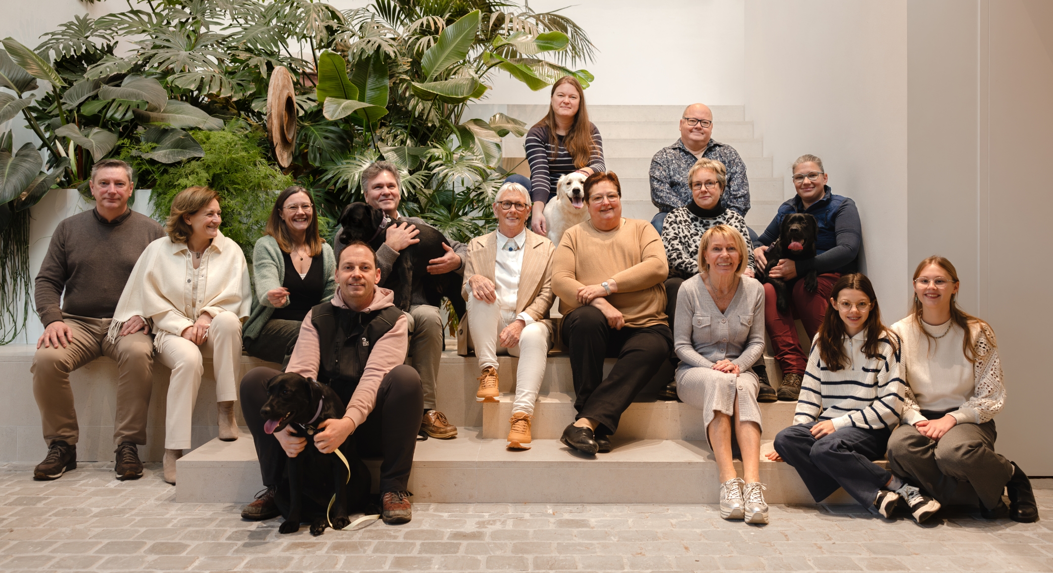 Groepsfoto met het hele team en vrijwilligers van Dogs for Blind. In totaal zijn ze met 13, bij hen zitten nog 3 zwarte en 1 witte honden. Ze zitten in een moderne traphal met witte muren, een brede trap uit roze beton en tropische planten op de achtergrond.