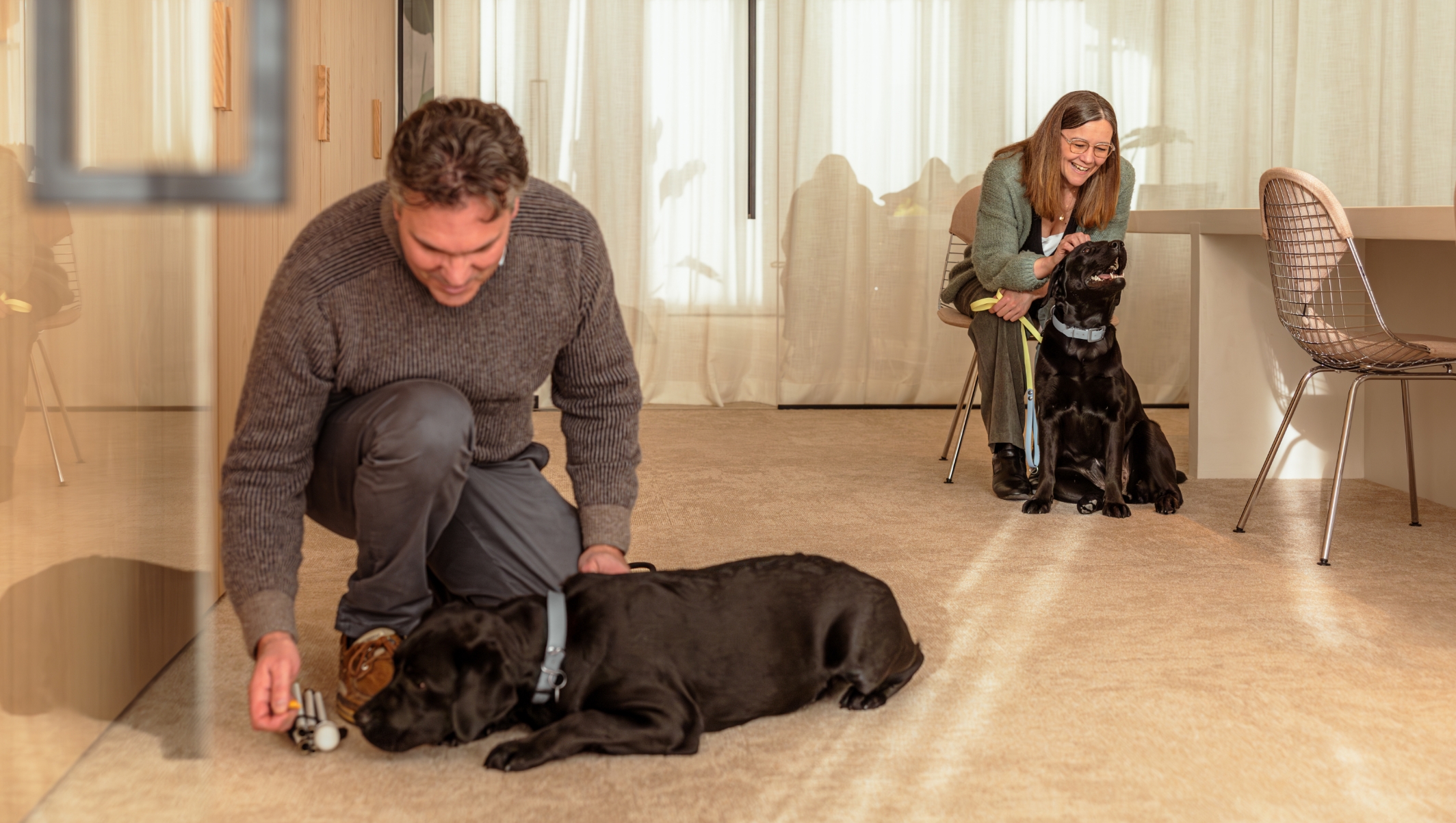 Afbeelding van Jo en Benedickt die samen in een ruimte zitten met elk een zwarte labrador. Ze genieten van het mooie moment samen. Benedickt zit links gehurkt en de hond ligt op de grond. Jo zit rechts op een bureaustoel en de hond zit neer voor haar.